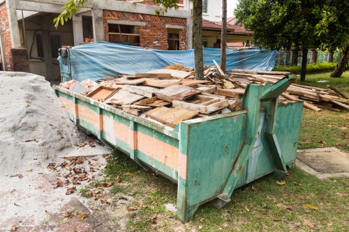 A large, industrial green metal skip positioned on a slightly uneven grassy area near a construction site in Highbury, North London. The skip is filled with a mixture of weathered, broken wooden planks, some with peeling paint and rough surfaces, and other construction debris. Behind the skip, there is a partially constructed brick building with exposed brickwork and wooden scaffolding, covered at the top with a blue tarpaulin. To the right, a lush green tree and a well-maintained lawn are visible, indicating a semi-residential or garden setting. The ground around the skip is speckled with fallen leaves and small debris, and the area appears to be in the process of renovation or clearance. The scene is well-lit with natural daylight, emphasizing the textures of the materials involved, aligning with rubbish removal services offered by Garden Clearance Highbury in the local North London area.