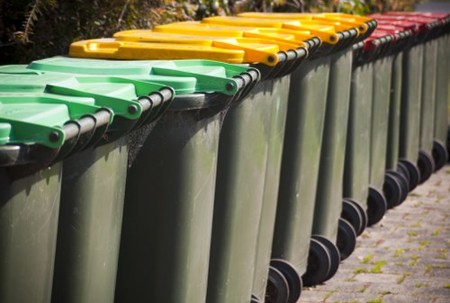 A row of large, wheeled outdoor rubbish bins lined up along a paved pathway, with the first bins featuring green lids, followed by yellow, and then red lids. The bins are made of durable plastic with a matte finish, and each has a black handle on the lid edge for opening. They are positioned close together, extending from the foreground to the background, with some slight overlapping. The pathway appears clean with minimal grass or moss growth between paving stones. The background features a blurred natural environment, possibly trees or shrubs, indicating this is an outdoor area near a residential or commercial property in Highbury. The image is well-lit, emphasizing the colours and textures of the bins and surroundings. Garden Clearance Highbury provides this type of refuse collection service, ensuring clutter-free spaces in the local area.