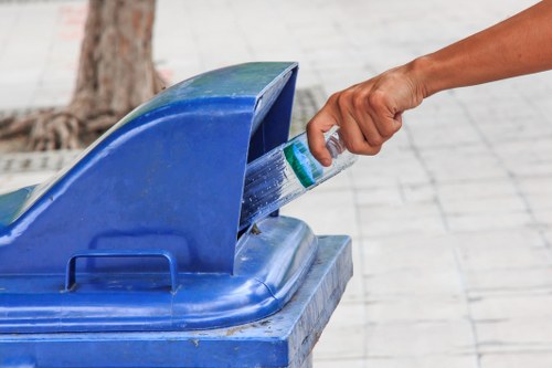 A person's hand is seen placing a small green and white container into a blue outdoor rubbish or recycling bin situated on a paved area. The bin has a rounded lid that is partially open, revealing some water or liquid inside. In the background, there is a tree trunk visible, indicating an outdoor setting, possibly in a garden or on a driveway. The surface around the bin is made of light grey paving stones, and the environment appears clean and well-maintained. This scene illustrates rubbish disposal or recycling practices, relevant to garden clearance services offered by Garden Clearance Highbury, serving areas such as Highbury and surrounding neighbourhoods in London, inferred from the context of the webpage.