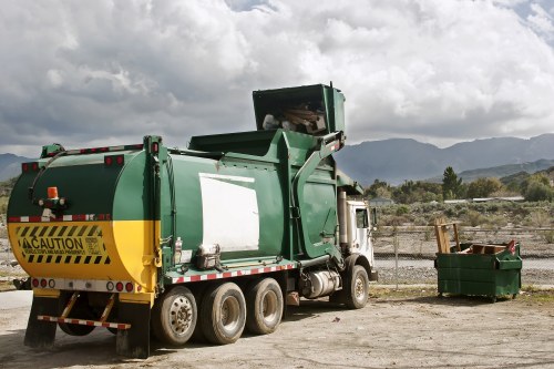 A large green and yellow waste collection truck, parked on a gravel surface near a chain-link fence, with mountains and a cloudy sky in the background. The truck, associated with rubbish removal services, features a prominent yellow caution stripe on the rear and an open hopper on top for waste loading. To the right of the truck, a green wheeled waste bin with an open lid is visible, containing miscellaneous rubbish or garden waste. The scene captures an outdoor setting typical for rubbish collection activities, with the truck positioned for waste loading or disposal. The subdued natural lighting emphasizes the utilitarian aspect of the vehicle and waste bins, aligning with the services offered by Garden Clearance Highbury for rubbish removal in the local area.