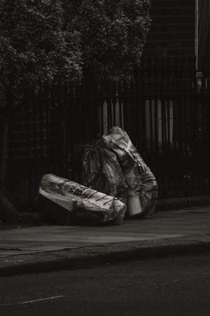 Two large black trash bags, made of thick plastic with visible creases and slight reflections, are positioned on the edge of a paved sidewalk in front of a dark metal fence with vertical bars. The bag