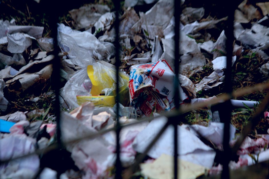 Two large black trash bags, made of thick plastic with visible creases and slight reflections, are positioned on the edge of a paved sidewalk in front of a dark metal fence with vertical bars. The bag