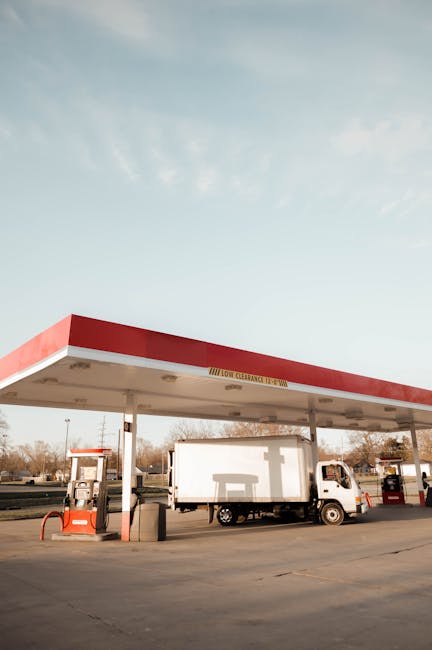 A small, white delivery truck is parked underneath a red and white canopy at a fuel station, with the front facing left. The truck is positioned close to two fuel pumps, one on each side, featuring bl
