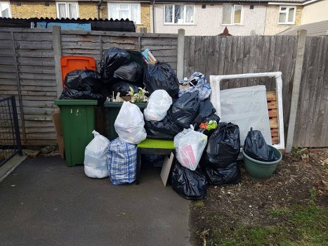 A collection of black and white rubbish bags filled with waste, piled on a small paved area next to a wooden fence. The bags are stacked alongside a green plastic wheelie bin, a large green garden planter, and an overturned white bed frame leaning against the fence. The waste appears to include general household rubbish, possibly garden debris, and packaging materials. The scene is outdoors in a residential setting, with neighbouring houses visible over the fence in the background. The overall appearance suggests a recent or ongoing rubbish clearance, typical of garden or property cleanup services offered by Garden Clearance Highbury in the Highbury area. The image reflects a typical scenario where professional rubbish removal is required to clear clutter and waste from a private outdoor space.