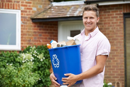 A young man with short light brown hair and a friendly smile is standing outdoors in front of a brick building, holding a blue recycling bin filled with various rubbish items including paper, plastic bottles, and packaging materials. The bin features a white recycling symbol on the side and appears to be made of durable plastic with a smooth finish. In the background, there is a white-framed window, some green foliage, and flowering plants suggesting a garden or residential property in Highbury, London. The man's attire includes a light pink short-sleeved shirt, and he is positioned on a paved or concrete outdoor area. Natural daylight illuminates the scene, capturing the outdoor environment where rubbish collection or garden clearance services by Garden Clearance Highbury might take place, emphasizing the importance of waste management in local residential settings.